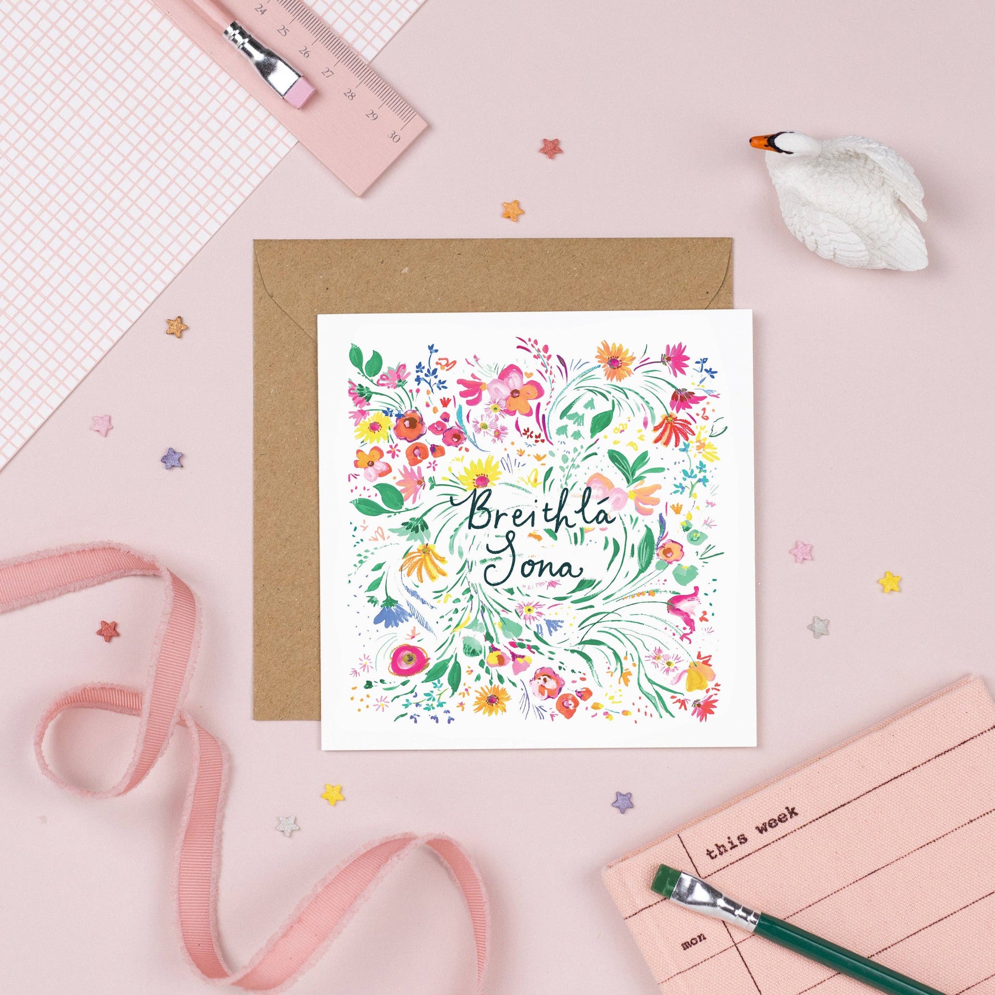 A greeting card with a floral design and the text 'Birthday bláthanna' in green, placed on a desk with a pink background, alongside a brown envelope and some decorative stars.