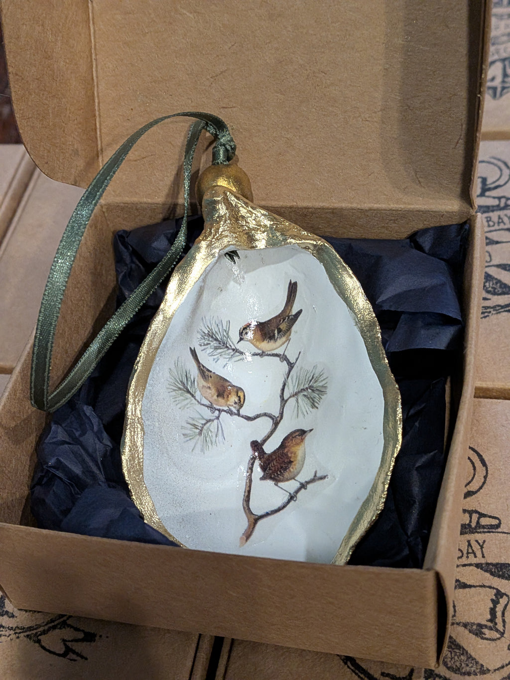 Decorative ornament with birds on a branch in a gold leaf frame, placed inside an open cardboard box.