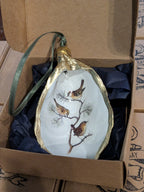 Decorative ornament with birds on a branch in a gold leaf frame, placed inside an open cardboard box.