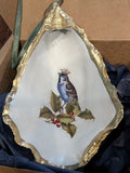 Decorative shell with a painted bird and holly design, gold rim, and green ribbon.