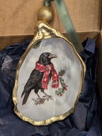 Decorative ornament with a crow wearing a red scarf and holly leaves, gold frame, and green ribbon.